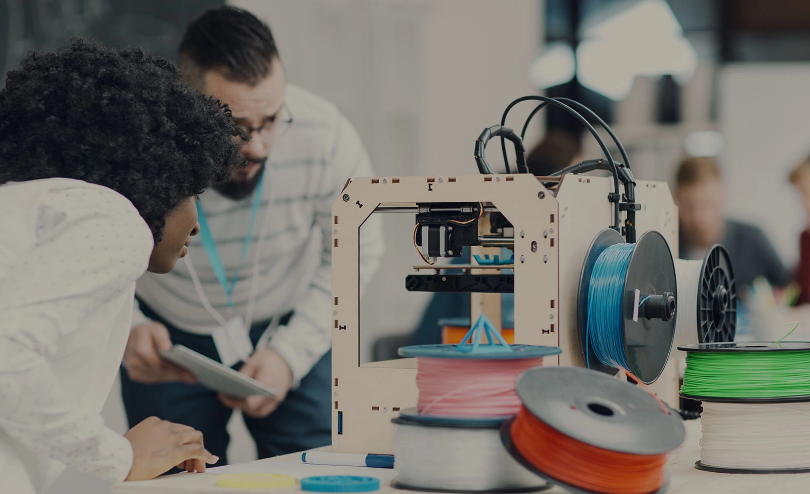 Diversity startup business team working together by 3D printer. Looking at printer working and discussing.  Man with beard and headphones around his neck holding digital tablet out and explaining to his african female coworker. Other coworkers working smoothly in background